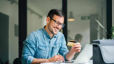 Happy man wearing a blue shirt and glasses holding a card and using buy now pay later services to purchase a product on his office computer