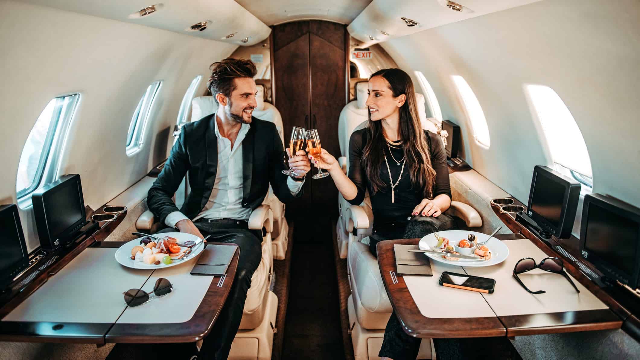 a couple clink champagne glasses on board a private aircraft with gourmet food plates set in front of them. They are wearing designer clothes and looking wealthy.