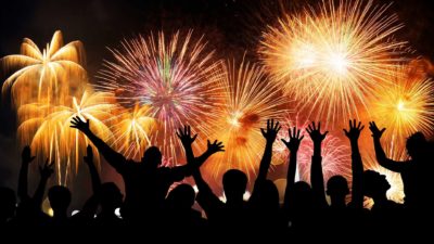 a group of people in shadow profile leap and hold their arms high in wonder of a fireworks display that fills the sky with light and colour and spectacular shapes.