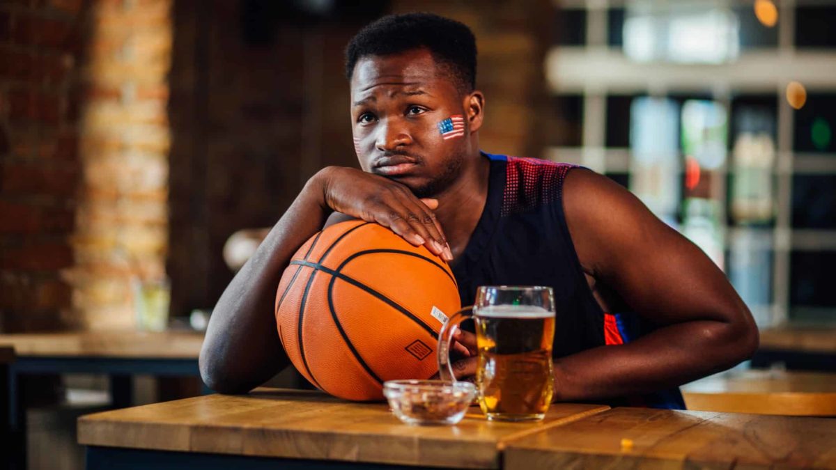 a man sits at a bar leaning sadly on his basketball wearing a US flag sticker on his cheekbone near a half drunk beer and looking despondent as though his basketball team has just lost a game.