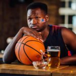 A man sits at a bar leaning sadly on his basketball.