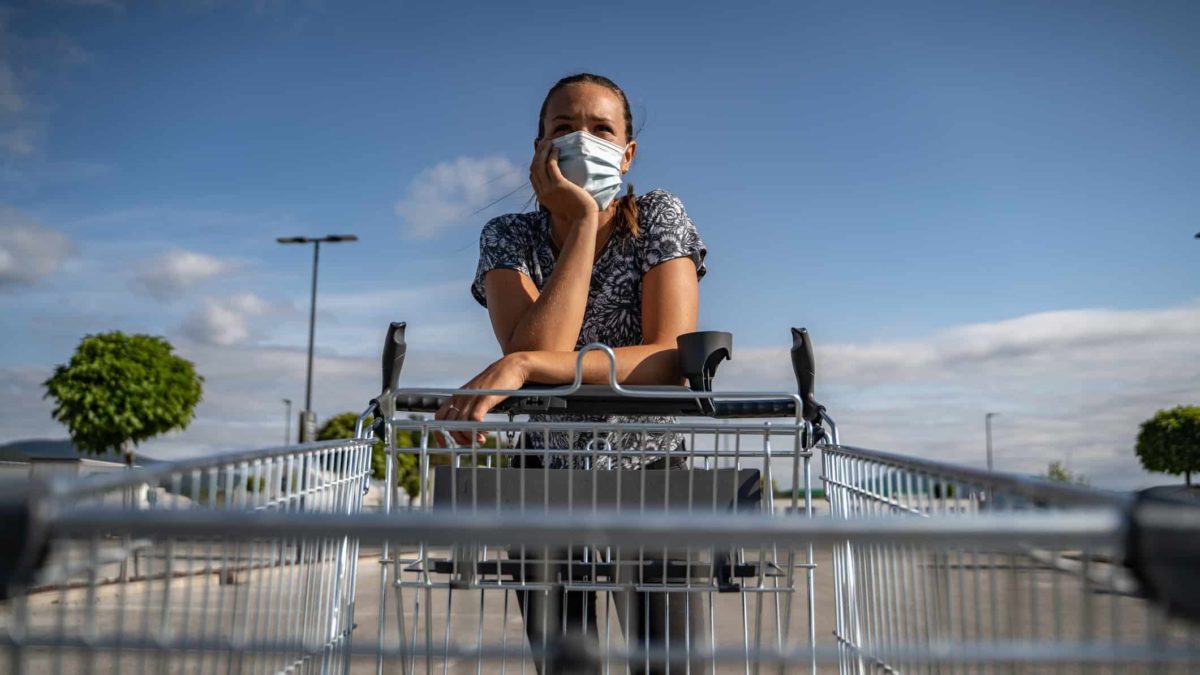 A frustrated woman wearing a COVID-19 mask leans over an empty supermarket shopping trolley