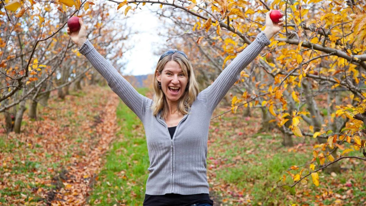 a girl stands in an apple orchard holding two red apples in raised arms with a happy, celebratory look on her face with a large smile and a pretty country background to the picture.