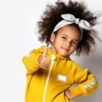 A happy girl in a yellow playsuit with a zip gives the thumbs up.