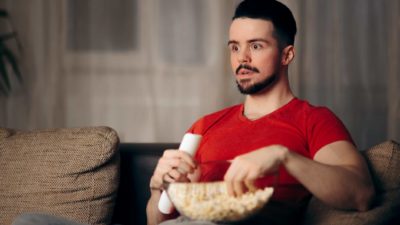 a man sits mesmerised by his television holding the remote in one hand with the other in a large bowl of popcorn as he sits in his lounge room and watched the TV intently.
