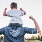 A boy sits on his dad's shoulders, both are flexing their biceps in unison.