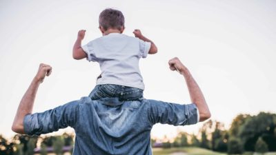 A boy sits on his dad's shoulders, both are flexing their biceps in unison.