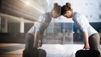 A businesswoman holding a briefcase rests her head against the glass wall of a city building, she's not having a good day.