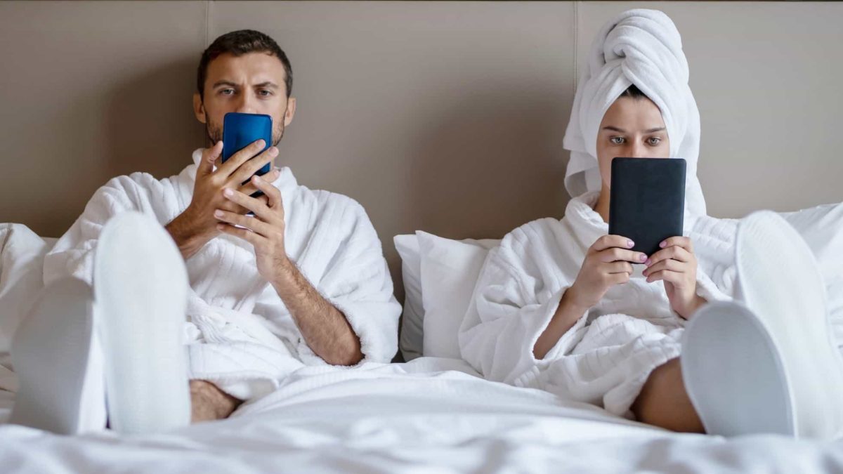 A couple sits on the bed in their hotel room wearing white robes, both have seen the bad news on their phones.