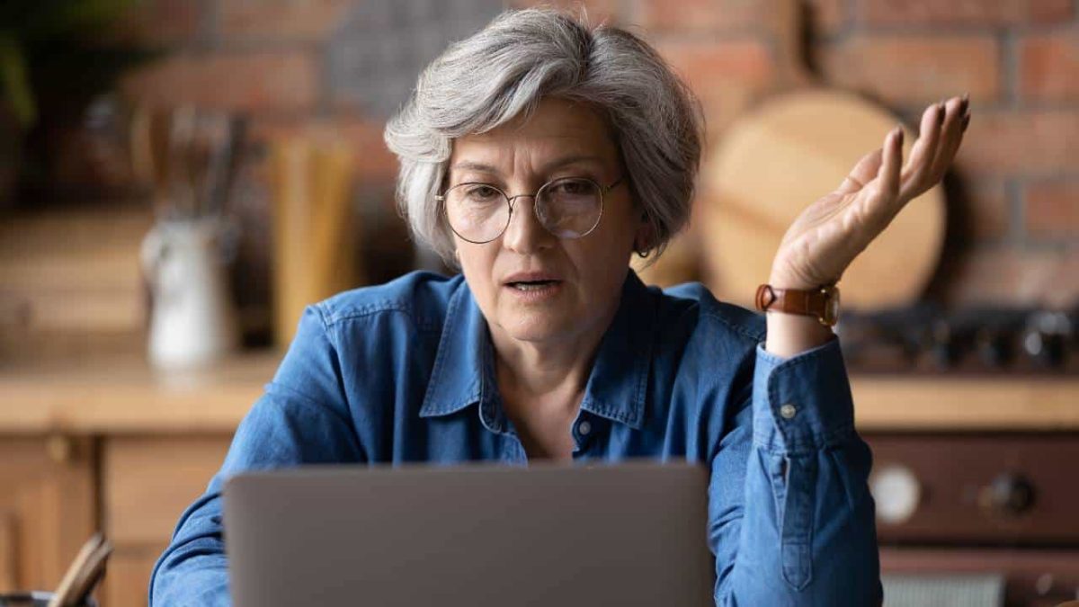 An older woman with grey hair and wearing glasses looks at her laptop screen with her hand outstretched to demonstrate that she doesn't understand what she is reading