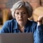 An older woman with grey hair and wearing glasses looks at her laptop screen with her hand outstretched to demonstrate that she doesn't understand what she is reading