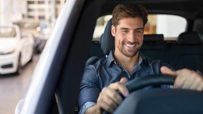 A handsome smiling man sits in the front seat of an electric vehicle with his hands on the wheel feeling pleased that the Carsales share price is going up and the company will shortly pay its biggest dividend ever