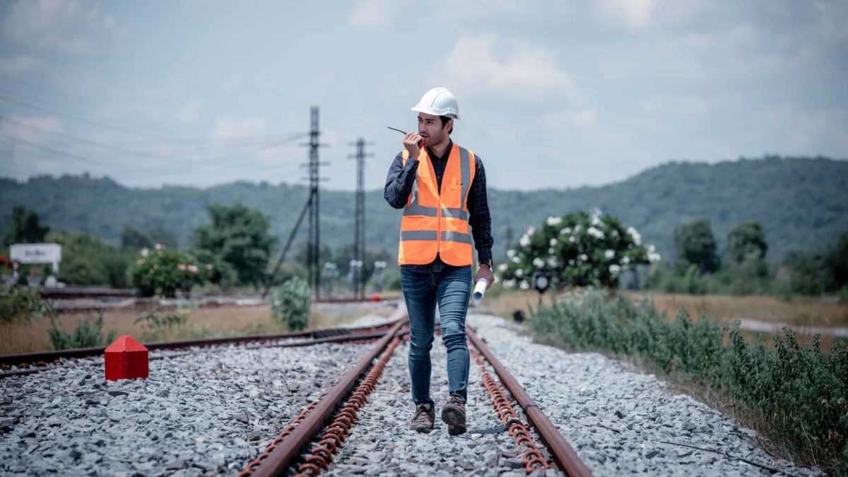 A railway worker walks along the train tracks in a visi vest and speaking into a walkie talkie.