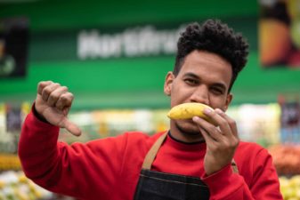 a supermarket employee holds an upside down banana in front of his mouth and his thumbs down as if showing his disapproval of something.