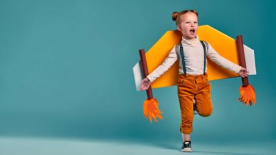 A little girl with red hair runs excitedly with a rocket strapped to her back, trying to launch.