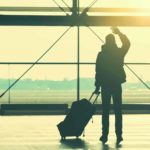 A person holding a suitcase waves goodbye as the sun sets outside the airport terminal.