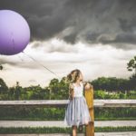 A girl stands at a wooden fence holding a big, inflated balloon looking at dark clouds looming ominously behind her.