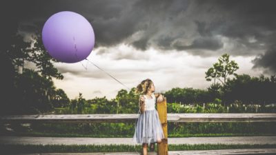 A girl stands at a wooden fence holding a big, inflated balloon looking at dark clouds looming ominously behind her.