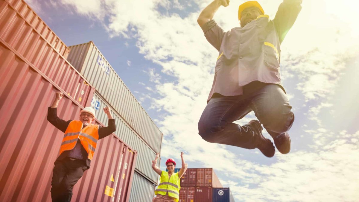 Workers at the port joyfully jump high in the air with shipping containers in the background.