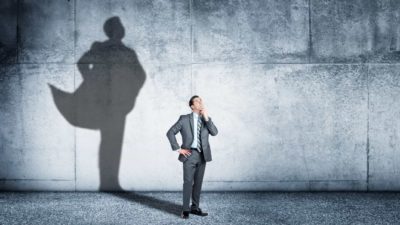 A man stands pondering the future while his shadow on the wall behind reveals he is wearing a cape.