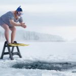 A man stands on a ladder in a stripey one-piece swimsuit, ready to plunge into the freezing water through a hole in the ice.