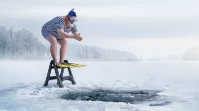 A man stands on a ladder in a stripey one-piece swimsuit, ready to plunge into the freezing water through a hole in the ice.