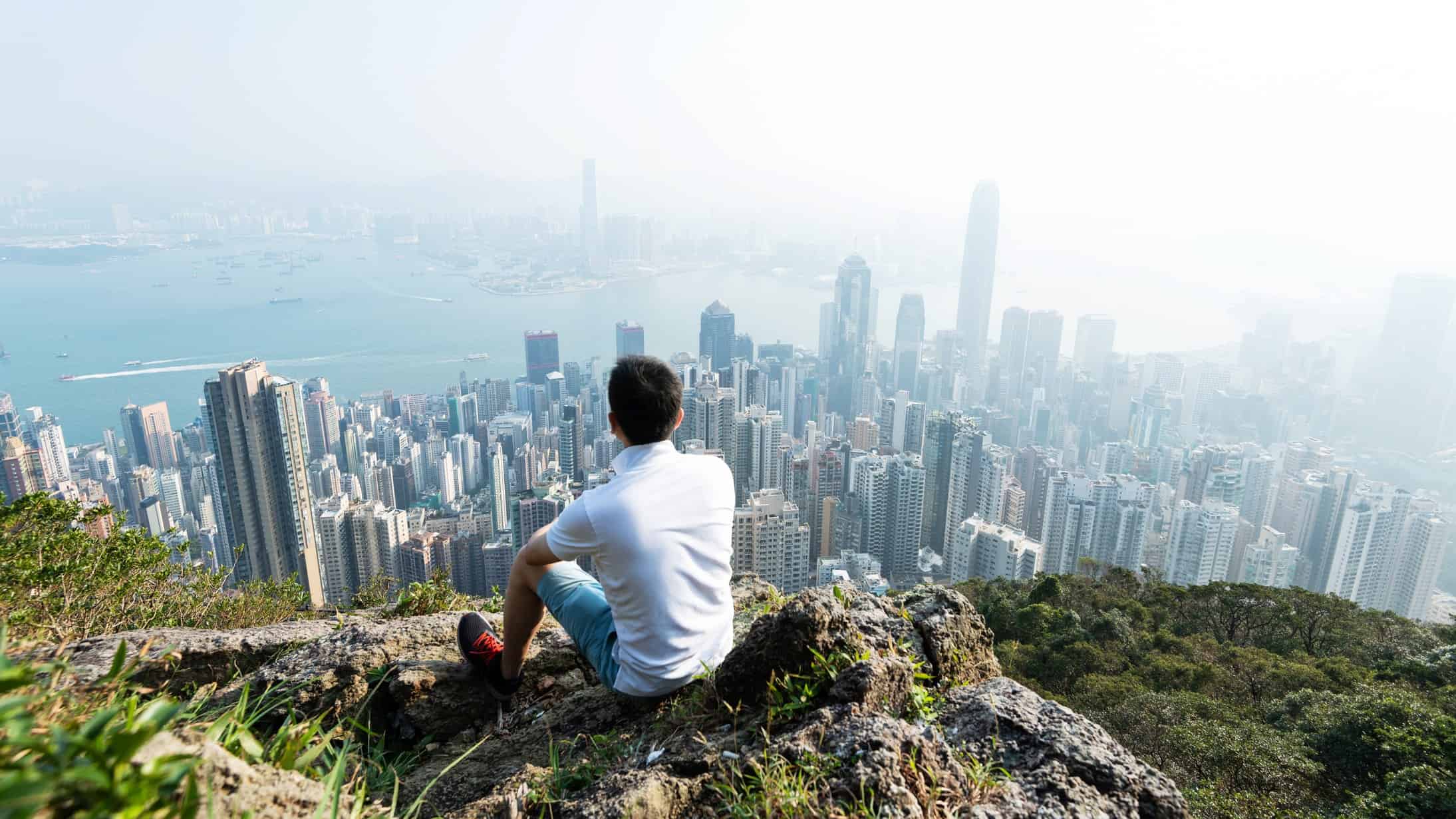 a man sits on a ridge high above a large city full of high rise buildings as though he is thinking, contemplating the vista below.
