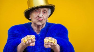An older female ASX investor holds a gangster-style fist pump pose showing off gold rings with dollar signs on them.