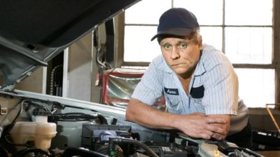 A mechanic rests his arms on a car he's working on, looking under the bonnet with a glum look on his face..