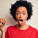 A woman holds a piece of pizza in one hand and has a shocked look on her face.