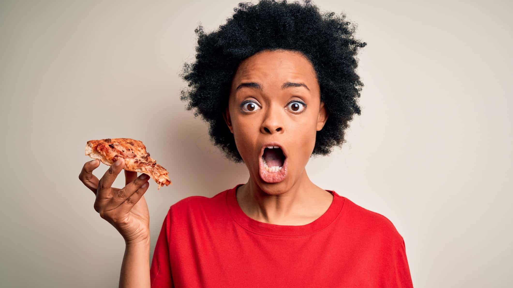 A woman holds a piece of pizza in one hand and has a shocked look on her face.