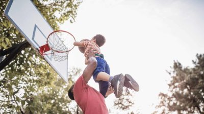 A dad holds his son up high so he can shoot the basketball into the ring.