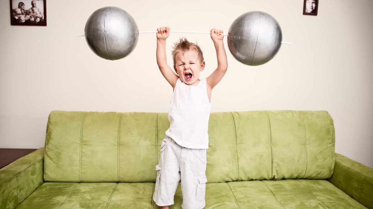 A little boy holds up a barbell with big silver weights at each end.