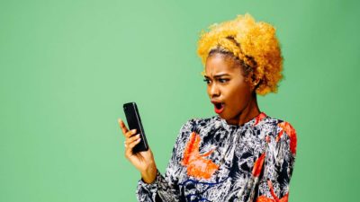 A woman with bright yellow hair wearing a brightly patterned blouse reacts to big news that she's reading on her phone.