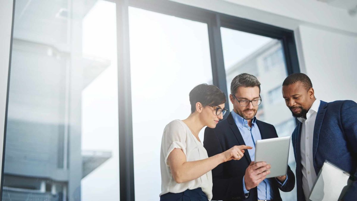 Three people in a corporate office pour over a tablet, ready to invest.