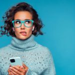 A woman wearing glasses has an uncertain look on her face as she bites her lip, she's just read some news on her phone.