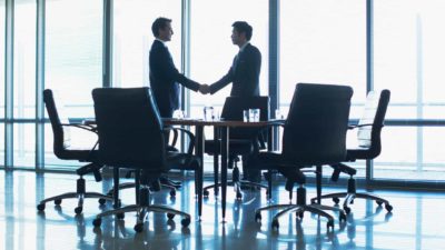 A silhouette shot of two business man shake hands in a boardroom setting with light coming from full length glass windows beyond them.