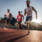Three male athletes sprint on an athletics track with the sun low on the horizon behind them representing the race between ASX lithium shares to outperform