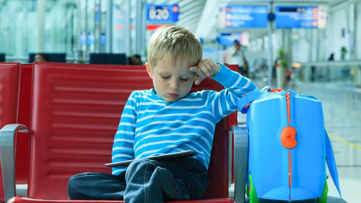 a small boy sits alone with his brightly coloured suitcase next to him in a deserted airport while he rests a hand against his head and looks down into his lap as though he is weary.