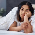 a young woman props her hand under the face as she pokes her head out from under a luxurious doona in a bedroom decorated with flowers and a stylish lamp.