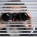a business man in a suit holds binoculars to his eyes and pokes them through old fashioned venetian blinds.