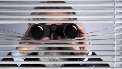 a business man in a suit holds binoculars to his eyes and pokes them through old fashioned venetian blinds.