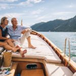A couple sit on the deck of a yacht with a beautiful mountain and lake backdrop enjoying the fruits of their long-term ASX shares and dividend income.