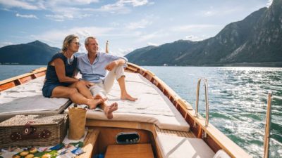 A couple sit on the deck of a yacht with a beautiful mountain and lake backdrop enjoying the fruits of their long-term ASX shares and dividend income.