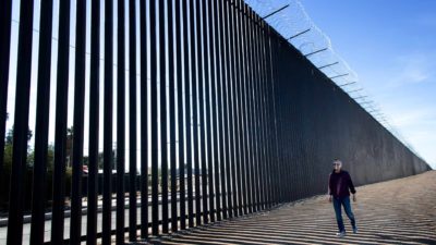 a man walks along the ground besidea high border fence topped with barbed wire.