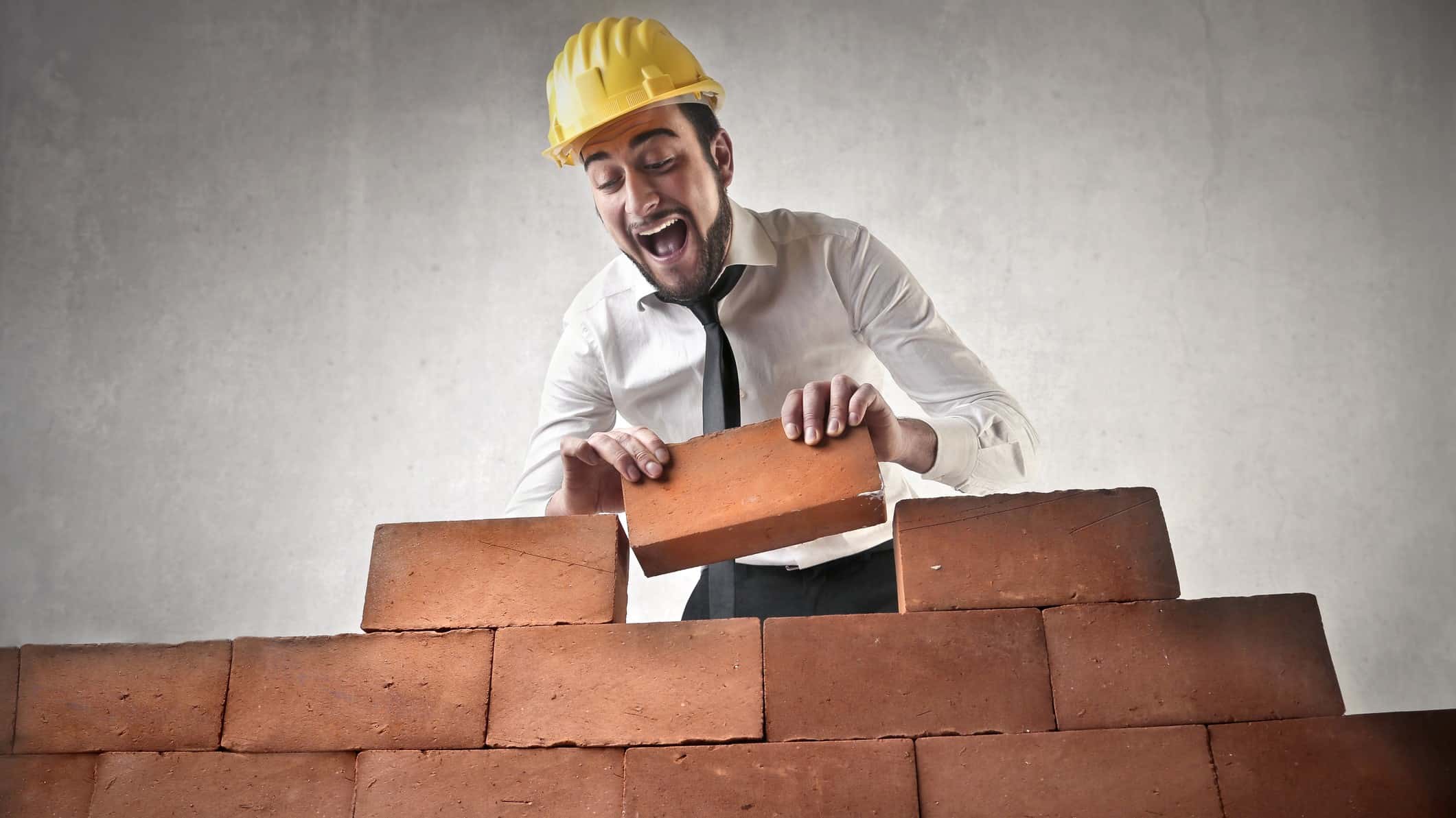 a man wearing a hard hat, a shirt and a tie, lays a brick on a wall he is building with a look of happy joy on his face.