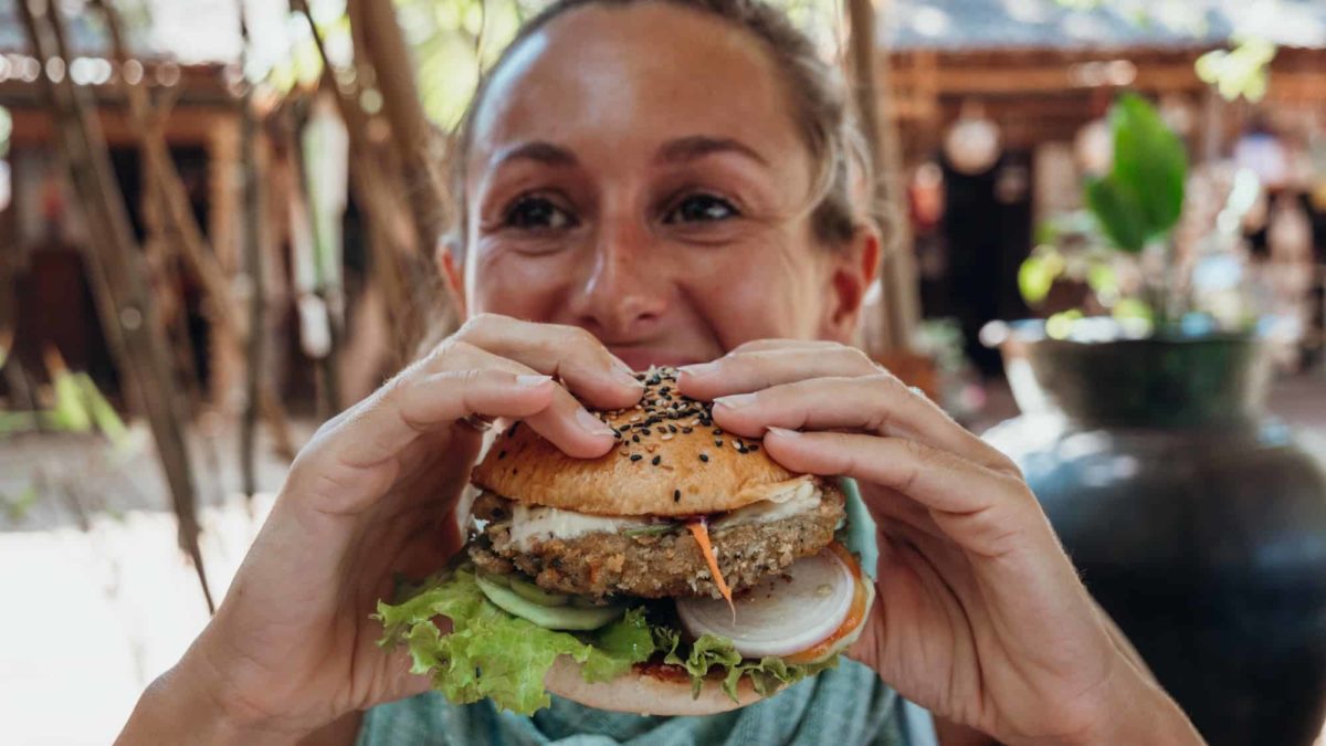 A woman smiles as she holds up a vegan burger to her mouth.