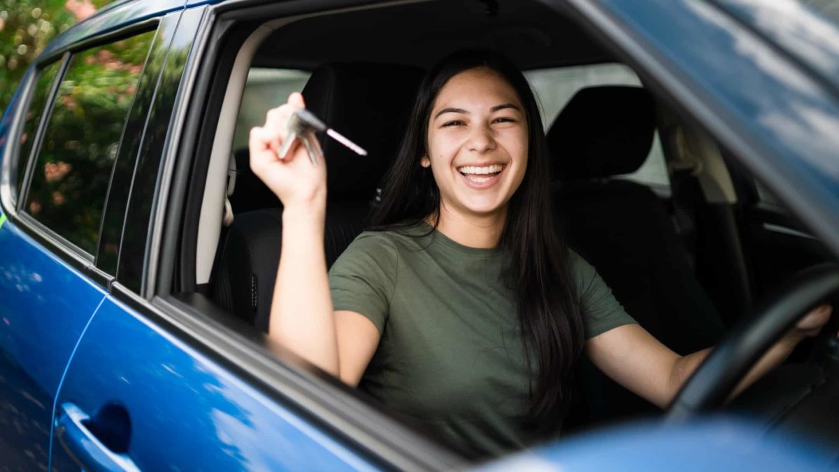 a young woman smiles widely as she holds up the keys while sitting in the driver's seat of her new car.