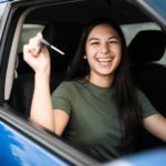 a young woman smiles widely as she holds up the keys while sitting in the driver's seat of her new car.