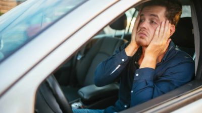 A man holds his hands to the sides of his face and pulls it down in despair as he sits at the wheel of a car that is not moving, as though in a traffic jam.